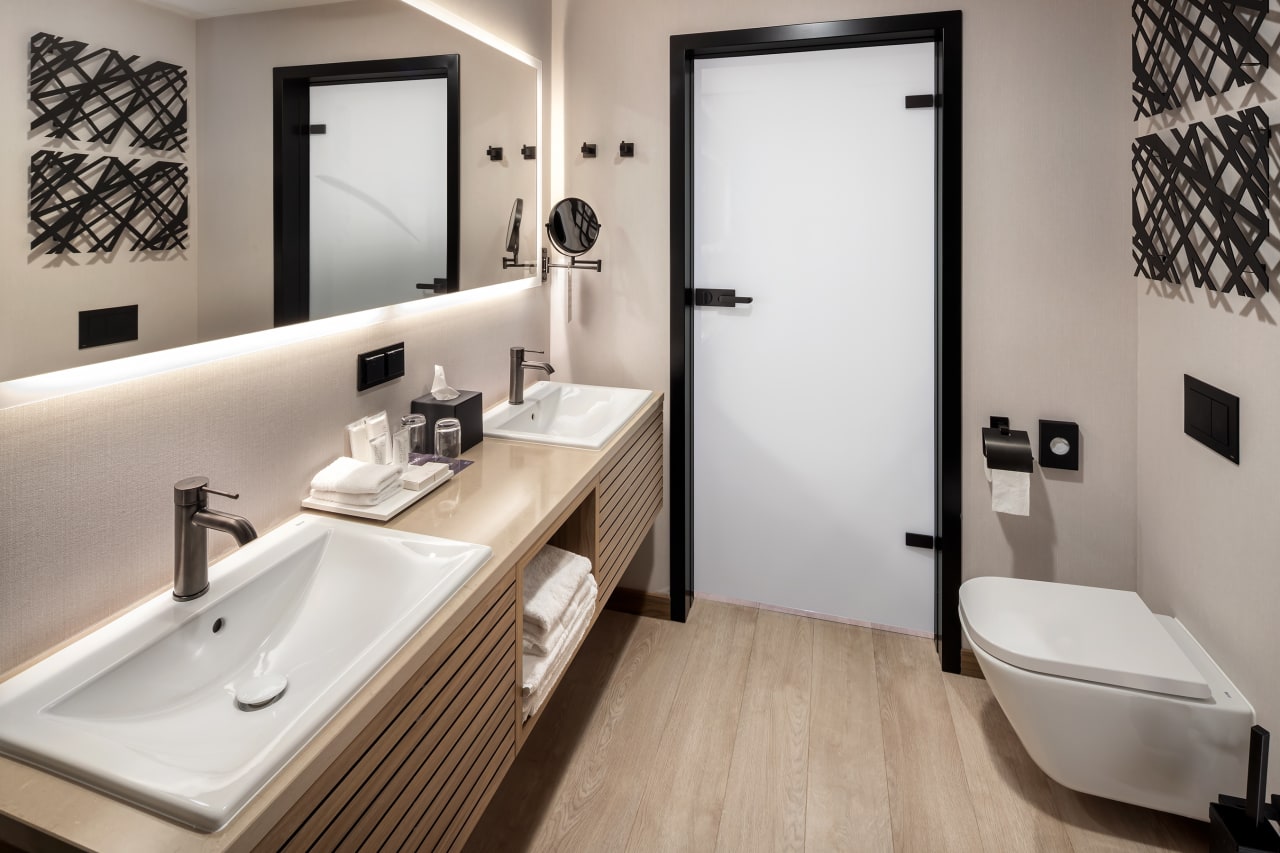 El interiorismo, obra de Tremend Studio, presenta un diseño armonioso caracterizado por el uso de tonos cálidos y naturales con detalles en madera que crean un ambiente agradable y relajado. Los baños de las habitaciones cuentan con confortables bañeras encastradas o duchas walk-in y mamparas de cristal, que generan una sensación de amplitud y espaciosidad. La atmósfera exclusiva y acogedora de estos baños se logra gracias a los muebles con acabados en madera, los lavabos Diverta sobre encimera y los inodoros The Gap Rimless. Por otro lado, los baños públicos también destacan por su minimalismo, con una planta diáfana en la que cobran protagonismo los lavabos Diverta y los urinarios Hall. Los espejos con iluminación LED integrada realzan el espacio con un suave resplandor y añaden al mismo tiempo un toque de sofisticación.4