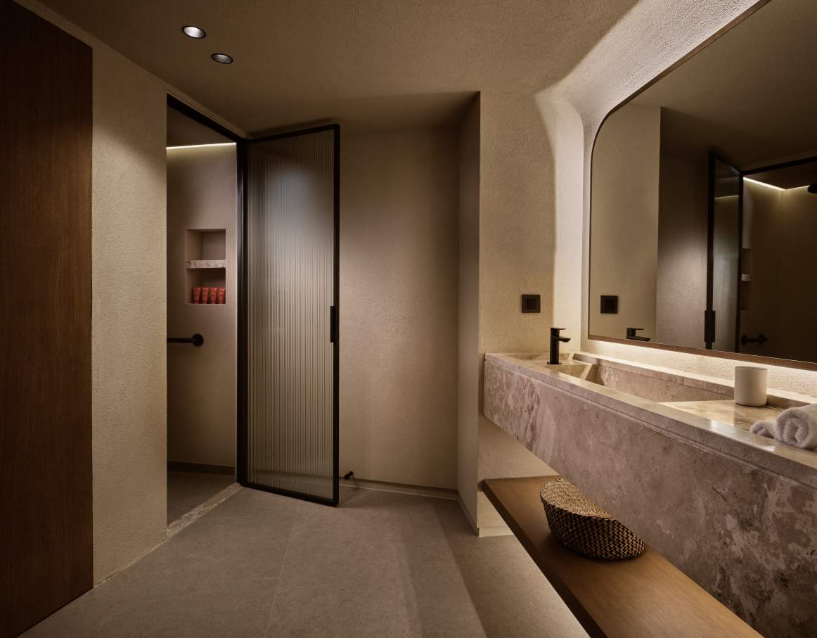 El interior de este complejo hotelero presenta una estética minimalista en tonos tierra, materiales de primera calidad y una iluminación tenue para crear una atmósfera relajante, propia de un spa. Los baños cuentan con una amplia encimera suspendida de mármol y un sobrio lavabo integrado, complementado con la elegante grifería Cala en negro mate, que crea un llamativo contraste con los tonos más claros del mármol y las paredes.