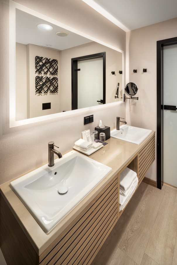 El interiorismo, obra de Tremend Studio, presenta un diseño armonioso caracterizado por el uso de tonos cálidos y naturales con detalles en madera que crean un ambiente agradable y relajado. Los baños de las habitaciones cuentan con confortables bañeras encastradas o duchas walk-in y mamparas de cristal, que generan una sensación de amplitud y espaciosidad. La atmósfera exclusiva y acogedora de estos baños se logra gracias a los muebles con acabados en madera, los lavabos Diverta sobre encimera y los inodoros The Gap Rimless. Por otro lado, los baños públicos también destacan por su minimalismo, con una planta diáfana en la que cobran protagonismo los lavabos Diverta y los urinarios Hall. Los espejos con iluminación LED integrada realzan el espacio con un suave resplandor y añaden al mismo tiempo un toque de sofisticación.8