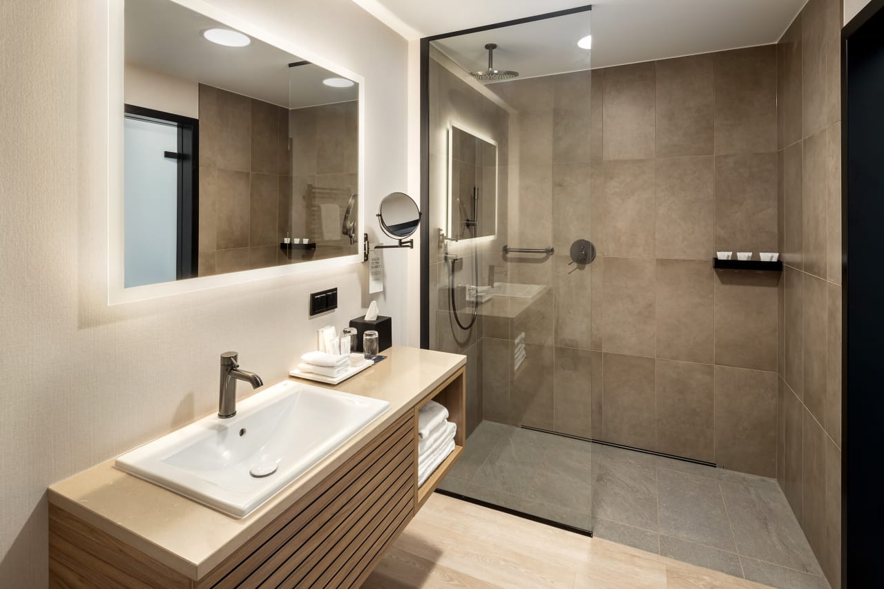 El interiorismo, obra de Tremend Studio, presenta un diseño armonioso caracterizado por el uso de tonos cálidos y naturales con detalles en madera que crean un ambiente agradable y relajado. Los baños de las habitaciones cuentan con confortables bañeras encastradas o duchas walk-in y mamparas de cristal, que generan una sensación de amplitud y espaciosidad. La atmósfera exclusiva y acogedora de estos baños se logra gracias a los muebles con acabados en madera, los lavabos Diverta sobre encimera y los inodoros The Gap Rimless. Por otro lado, los baños públicos también destacan por su minimalismo, con una planta diáfana en la que cobran protagonismo los lavabos Diverta y los urinarios Hall. Los espejos con iluminación LED integrada realzan el espacio con un suave resplandor y añaden al mismo tiempo un toque de sofisticación.1